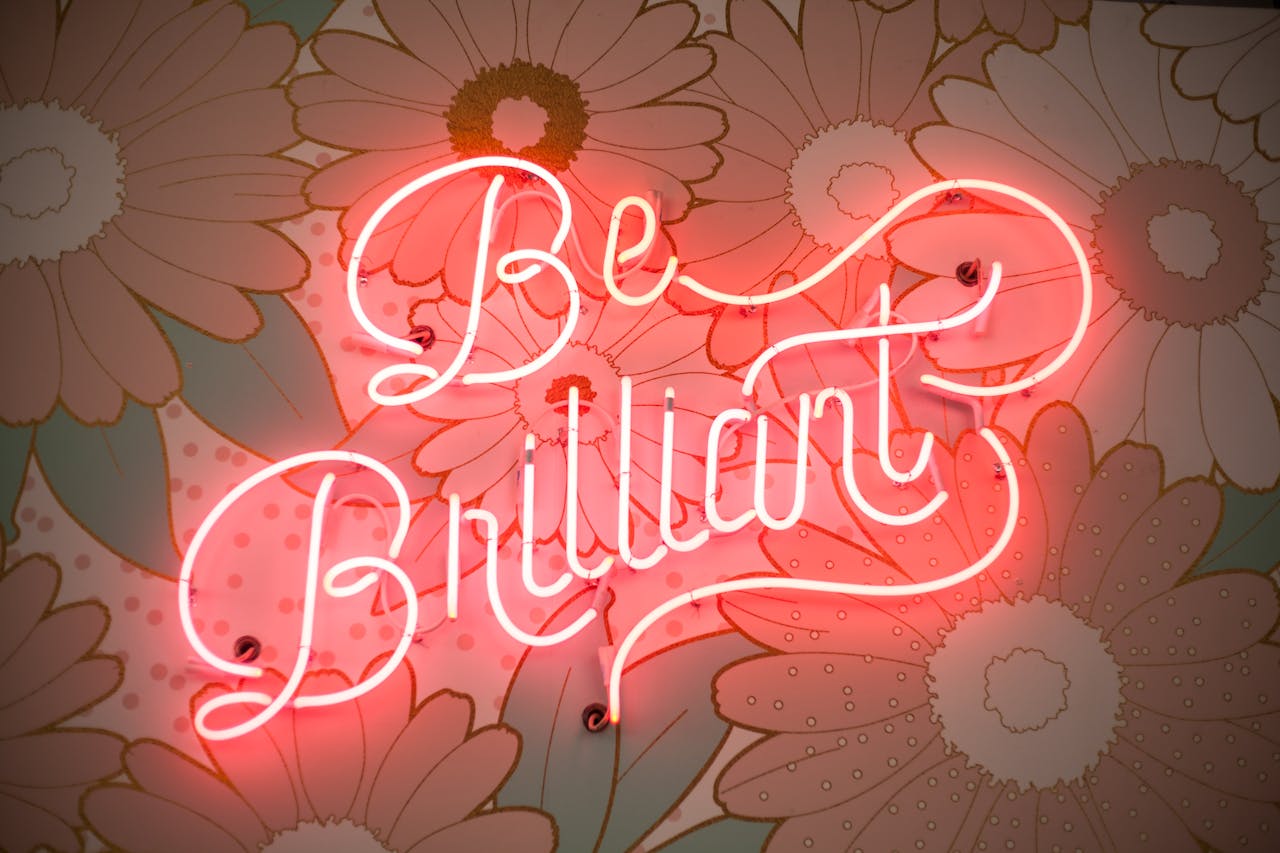 A glowing neon sign reading 'Be Brilliant' against a floral backdrop creates a vibrant and motivational atmosphere.